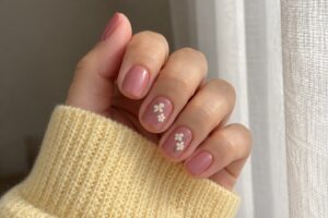 Short dusty rose nails with tiny white daisies, paired with a butter-yellow cardigan beside a sheer curtain.