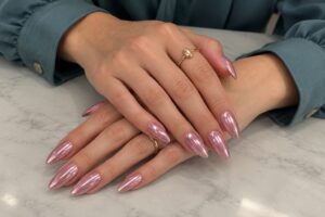 Dusty rose chrome almond nails on crossed hands with delicate gold rings and muted teal sleeves.