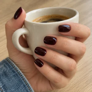 Glossy black cherry nails on a hand holding an ivory coffee cup