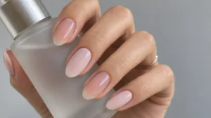 Sheer and milky pink almond nails on a hand holding a frosted glass bottle.