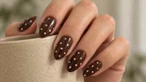 Chocolate brown almond nails with cream polka dots on a hand holding a ceramic cup.