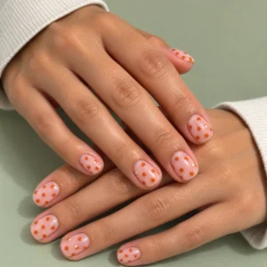 Close-up of short blush-pink nails with pumpkin-orange polka dots against a pale sage background.