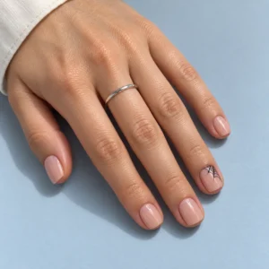 Short nude-pink nails with a delicate black spiderweb in one corner of the ring fingernail against a pale blue background.