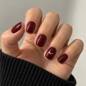 Close-up of short glossy burgundy nails with a small silver crescent moon on the ring fingernail.