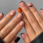 Close-up of short Halloween nails with black French tips, a tiny ghost, a black bat, and pumpkin-orange polish.