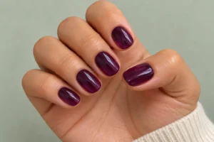 Close-up of short oval nails with glossy, translucent deep plum polish against a pale sage-gray background.