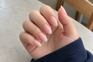 Close-up of sheer pink almond-shaped nails with a glossy finish and navy sleeve against a light tabletop.