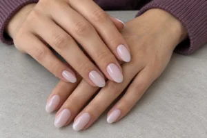 Close-up of overlapping hands with glossy milky nude almond nails and plum sleeves on a gray tabletop.