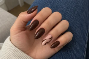 Close-up of glossy chocolate brown almond nails with a nude swirl accent nail against blue denim.