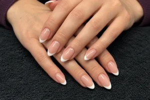 Two overlapping hands with glossy nude almond nails and white French tips resting on a charcoal towel.