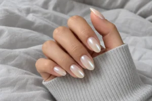 Close-up of milky ivory almond nails with a soft pearl sheen, framed by a gray sleeve against cotton bedding.