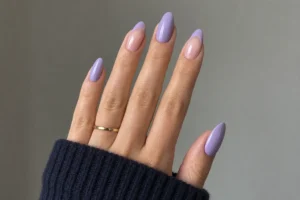 Close-up of muted lavender almond nails with nude and lavender French tip accents, a gold ring, and a navy sleeve.