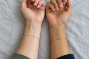 Two inner wrists with visible veins, one wearing a silver chain bracelet and the other a gold chain bracelet, on gray fabric.