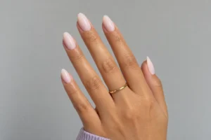 Close-up of milky pink almond nails with a thin gold ring and lavender sleeve against a light gray background.