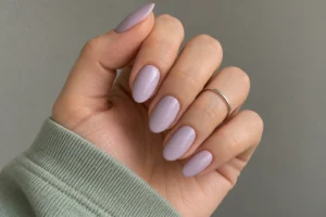 Close-up of glossy pale lilac almond nails with a silver ring and sage green sleeve.