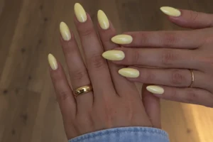 Two overlapping hands with glossy butter yellow almond nails and gold rings against a wooden floor.