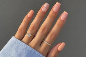 Close-up of peach cream square nails with peach French tips, white daisy accents, and gold rings.