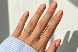 Close-up of glossy apricot coral oval nails with thin silver rings and a blue striped sleeve in sunlight.