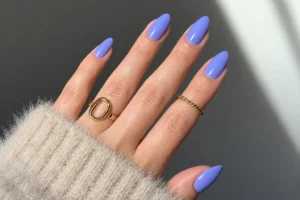 Close-up of glossy periwinkle almond nails with gold rings and an oatmeal fuzzy sleeve in natural sunlight.