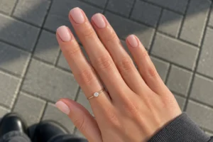 Close-up of glossy creamy beige squoval nails with a delicate gold ring above gray paving stones.