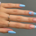 Spring manicure with pastel blue almond nails, blue French tips, nude accents, and delicate gold rings.