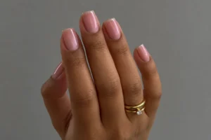 Close-up of sheer blush pink square nails with a subtle pearly sheen and stacked gold rings against a gray background.