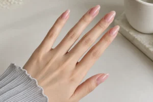 Close-up of glossy pink-beige almond nails with a gray ribbed sleeve beside a ceramic cup on a cream tabletop.