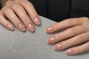 Two hands with glossy peachy pink oval nails and subtle ivory tips resting on a light gray cushion.