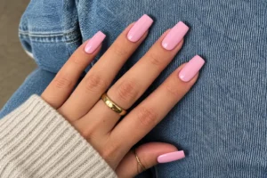 Close-up of glossy soft rose pink square nails with gold rings and an oatmeal sleeve resting on blue jeans.