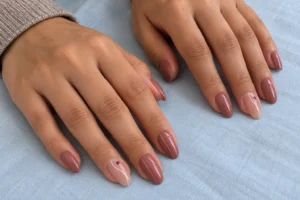 Rosy brown pink almond nails with fine ivory lines and burgundy dot accents resting on pale blue fabric.