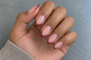 Close-up of almond nails with sheer pink bases and thin shimmering ruby red French tips.