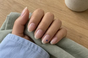 Close-up of sheer blush almond nails with one tiny blue star on the ring fingernail, resting on sage fabric.