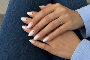Two overlapping hands with milky white almond nails and tiny red dots near the cuticles, resting on dark blue jeans.