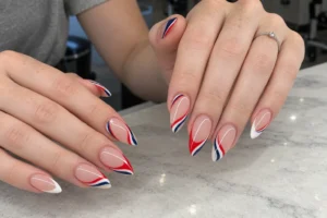 Close-up of nude almond nails with flowing red, navy blue, and white side lines above a gray marble table.
