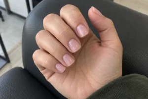 Close-up of sheer pink short square nails on a gently curled hand against a charcoal salon chair.