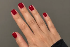 Close-up of glossy classic red short square nails on an outstretched hand against a warm gray background.