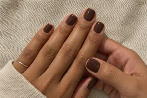Close-up of glossy chocolate brown short square nails on two hands against beige herringbone fabric.