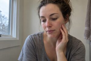 Fair-skinned woman gently touching her cheek beside a bathroom window in natural daylight.