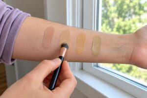 Four foundation swatches on a fair-skinned forearm being compared and blended beside a window in daylight.