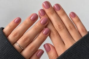 Short glossy dusty rose nails on two hands with charcoal knit sleeves.