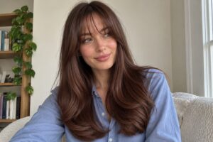 Woman with long chestnut brown layers and wispy bangs wearing a blue shirt.