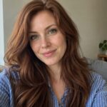 Woman with green eyes and long auburn hair wearing a blue striped shirt in natural daylight.