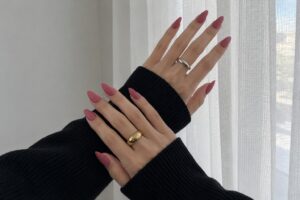 Matte dusty rose almond nails with black sweater sleeves and gold and silver rings beside sheer curtains.
